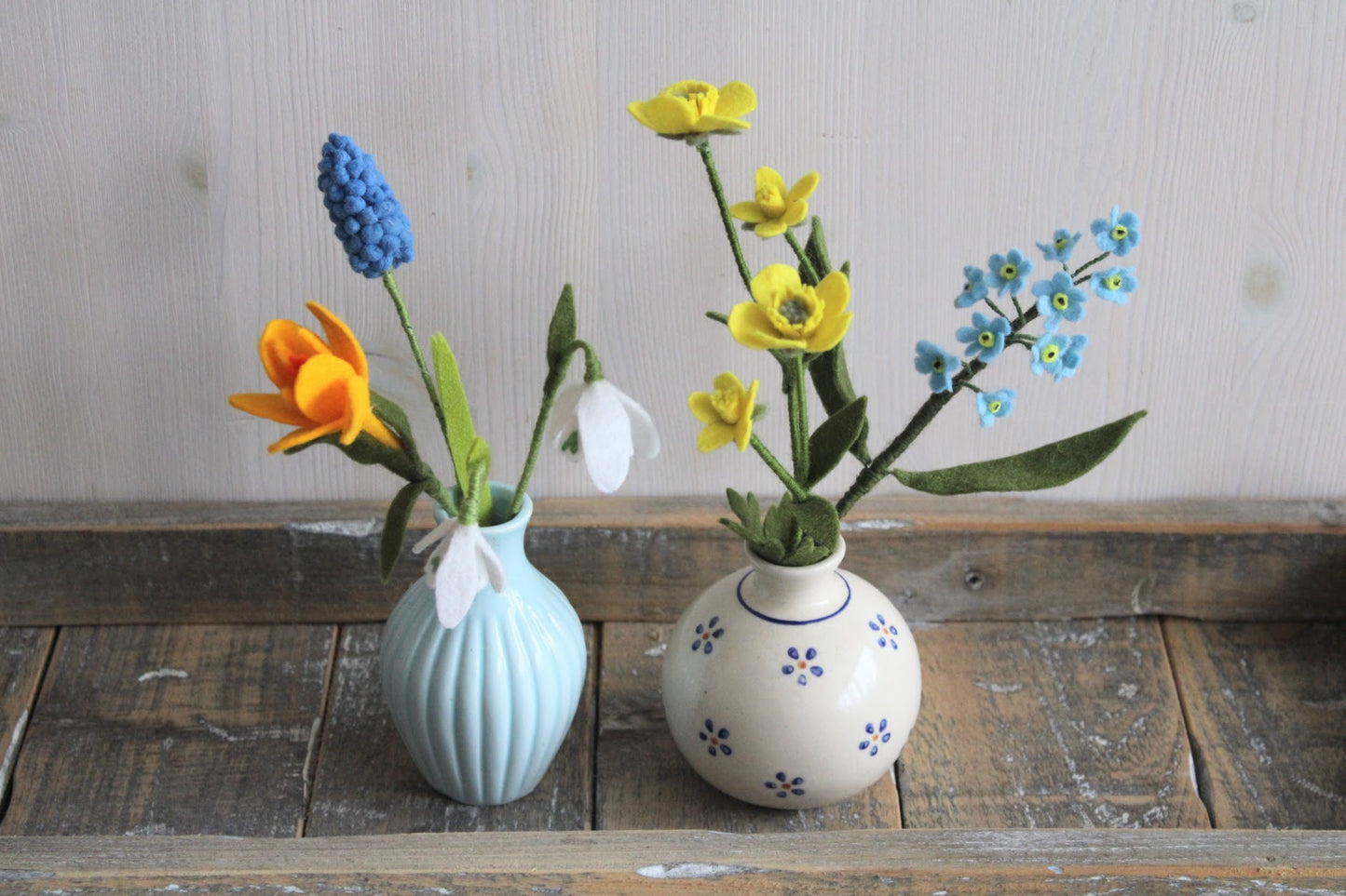 Handgemaakte viltbloemen gele krokus, sneeuwklokjes, blauw druifje, boterbloem, vergeet me nietjes verdeeld over twee vaasjes, blauw en met bloemetjes op een houten ondergrond