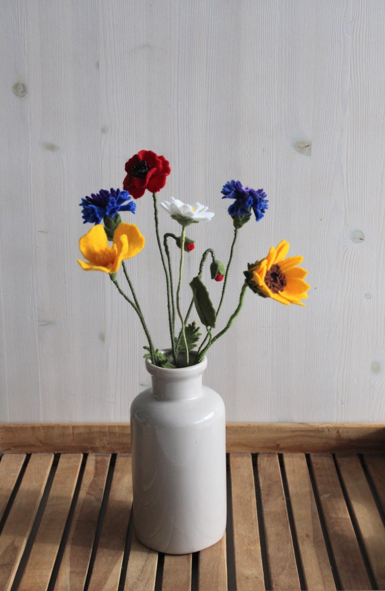 Handgemaakt viltbloemen veldboeket met geel slaapmutsje, margriet, korenbloemen, en klaproos in een grijs vaasje op een houten rustiek ondergrond