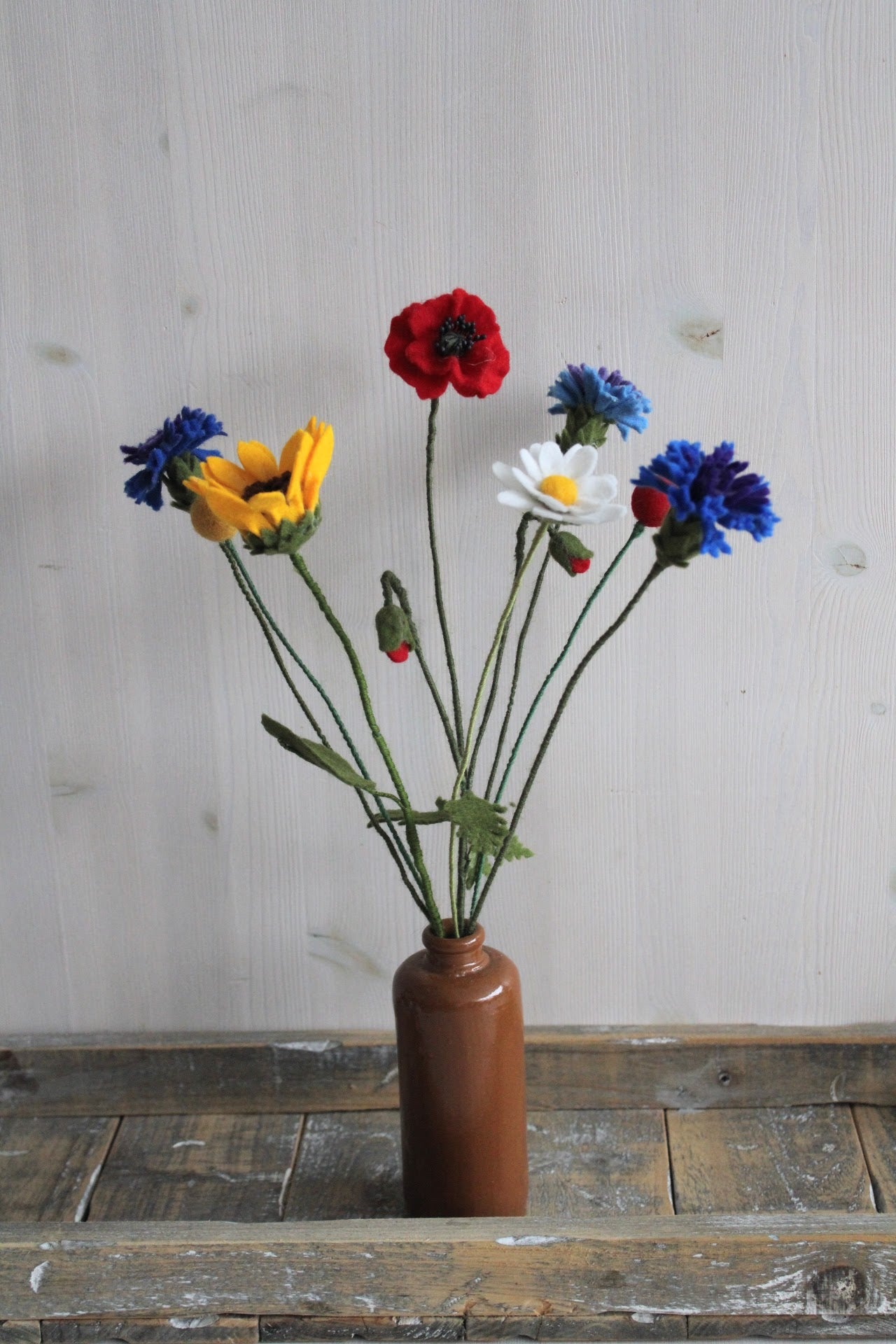 Handgemaakte vilten margriet, korenbloem, zonnebloem, klaproos en margriet op lange stelen bekleed met groene wol. Geplaatst in een bruine keramieken vaas op een rustieke houten ondergrond. Perfect als duurzame decoratie