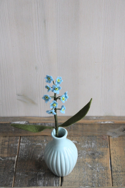 Handgemaakte vilten vergeet me nietje met blauwe bloemetjes en een geel hartje, op een korte steel bekleed met groene wol en twee blaadjes. Geplaatst in een blauw keramiek vaasje op een rustieke houten ondergrond. Perfect als duurzame decoratie of als onderdeel van een viltbloemenboeket