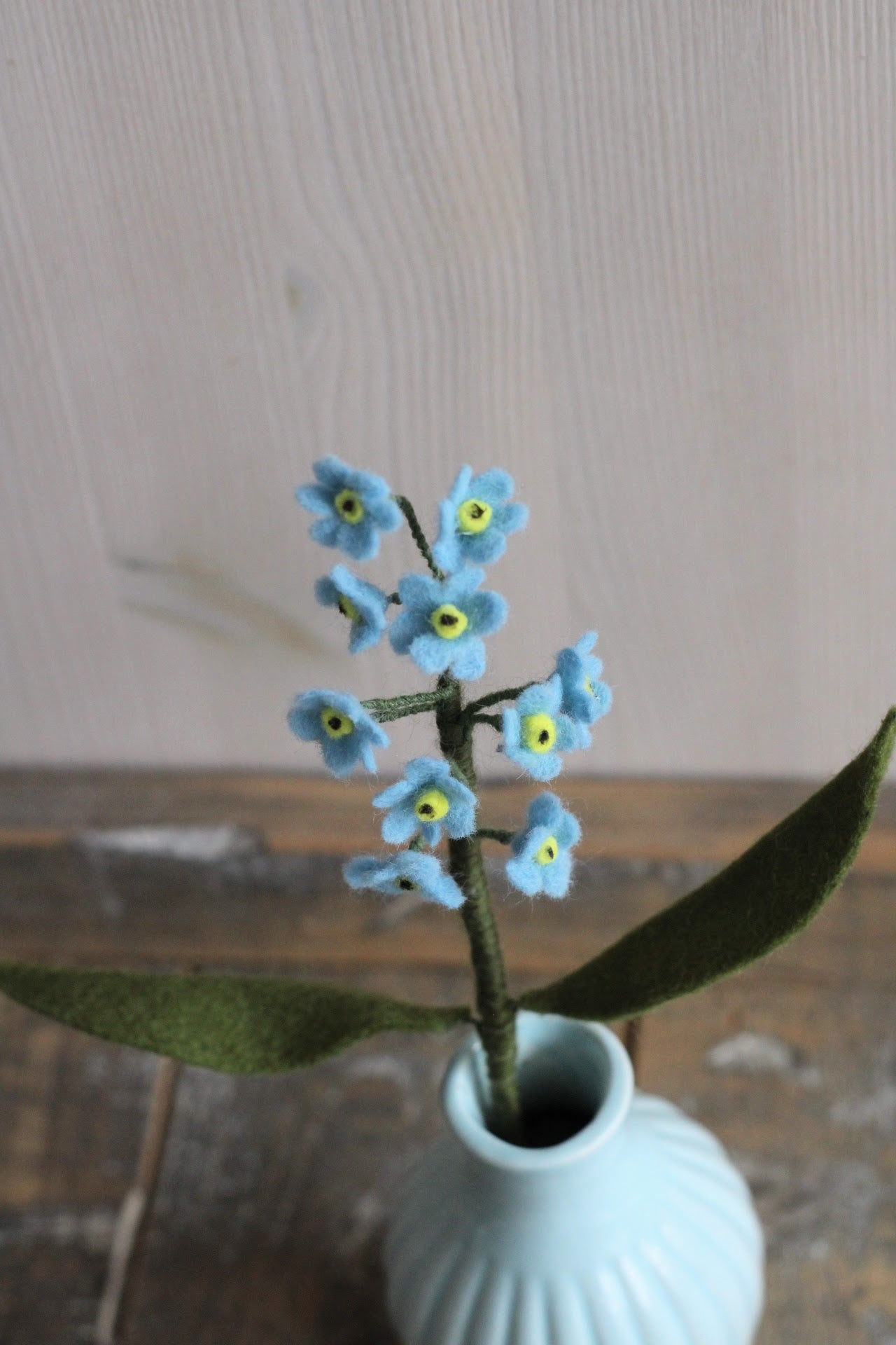 Handgemaakte vilten vergeet me nietje met blauwe bloemetjes en een geel hartje, op een korte steel bekleed met groene wol en twee blaadjes. Geplaatst in een blauw keramiek vaasje op een rustieke houten ondergrond. Perfect als duurzame decoratie of als onderdeel van een viltbloemenboeket