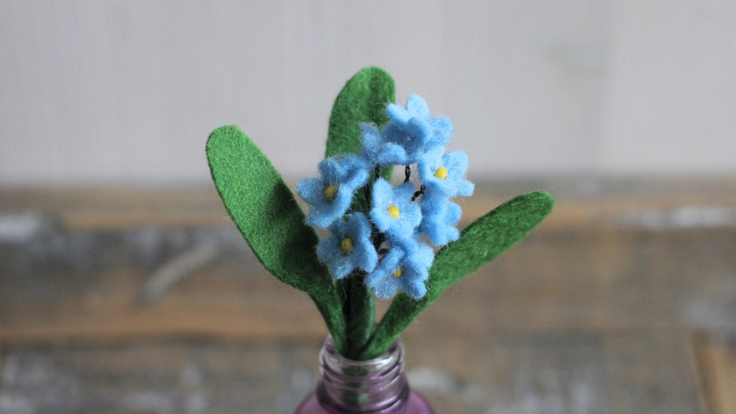Handgemaakte vilten vergeet me nietje met blauwe bloemetjes en een geel kralenhartje, op een korte steel bekleed met groene wol en twee blaadjes. Geplaatst in een paars glazen vaasje op een rustieke houten ondergrond. Perfect als duurzame decoratie of als onderdeel van een viltbloemenboeketje. hij kan ook perfect als broche gemaakt worden
