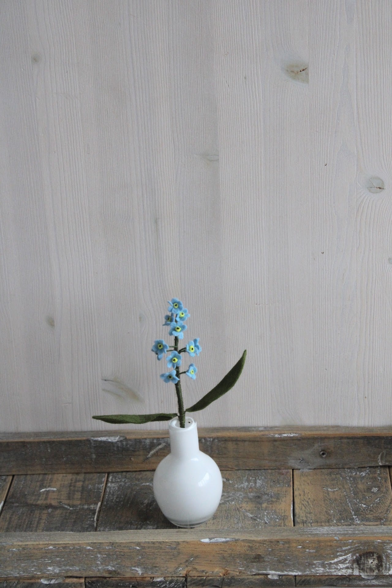 Handgemaakte vilten vergeet me nietje met blauwe bloemblaadjes en een geel hartje, op een korte steel bekleed met groene wol enn twee groen blaadjes. Geplaatst in een wit glazen vaasje op een rustieke houten ondergrond. Perfect als duurzame decoratie of als onderdeel van een viltbloemenboeket