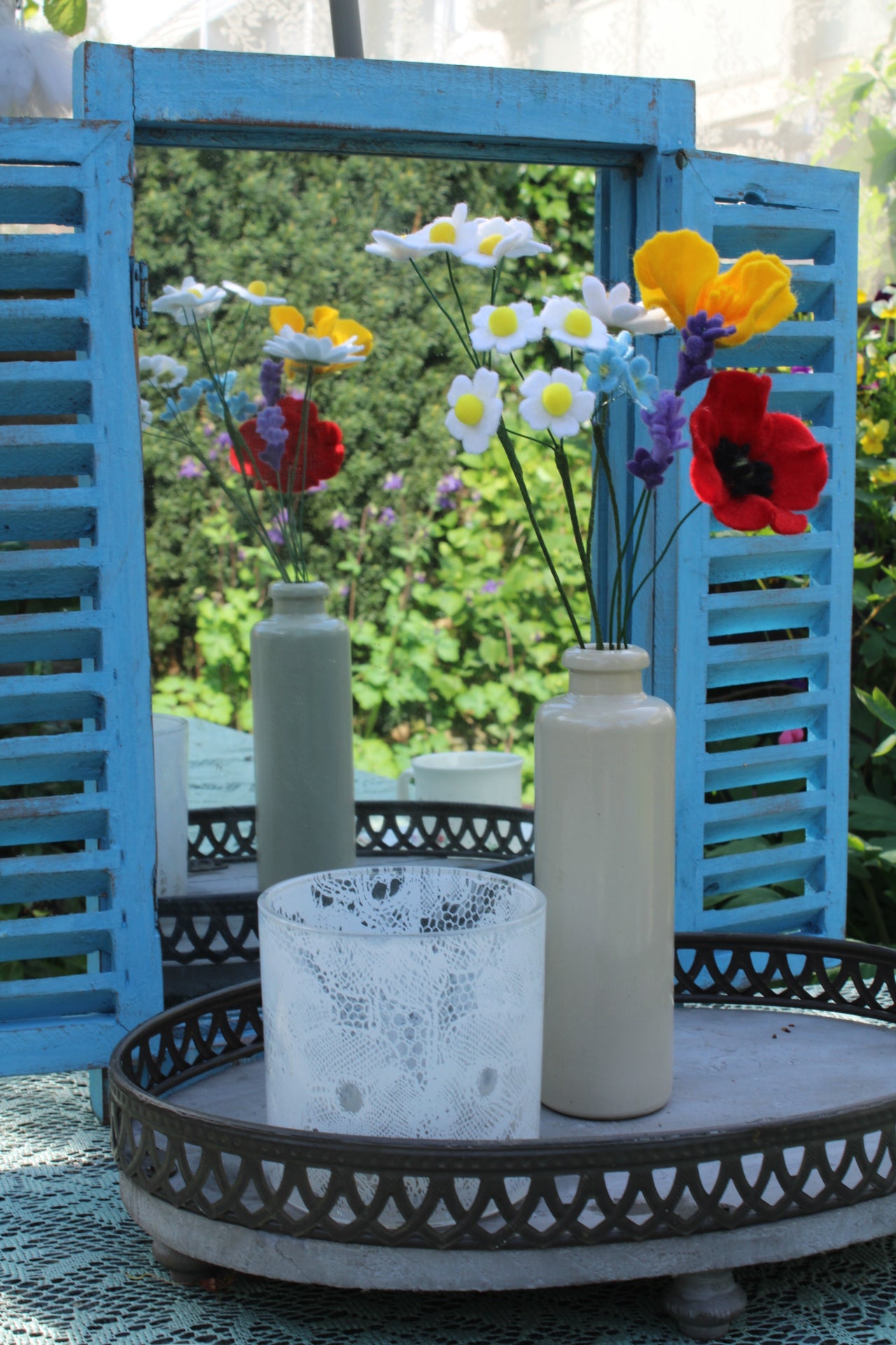 Handgemaakt boeket viltbloemen in een witte vaas, met madeliefjes, lavendel, vergeet-me-nietjes, gele en rode klaprozen, gepresenteerd voor een blauwe spiegel met tuin op de achtergrond
