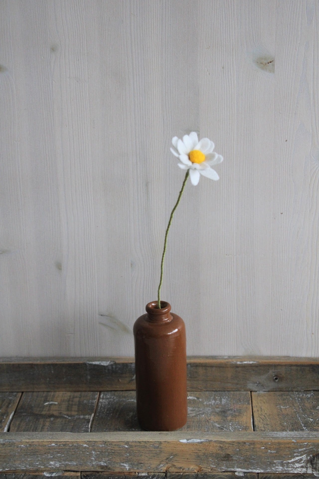 Handgemaakte vilten margriet met witte bloemblaadjes en een geel hart, op een lange steel bekleed met groene wol. Geplaatst in een bruine keramieken vaas op een rustieke houten ondergrond. Perfect als duurzame decoratie of als onderdeel van een viltbloemenboeket