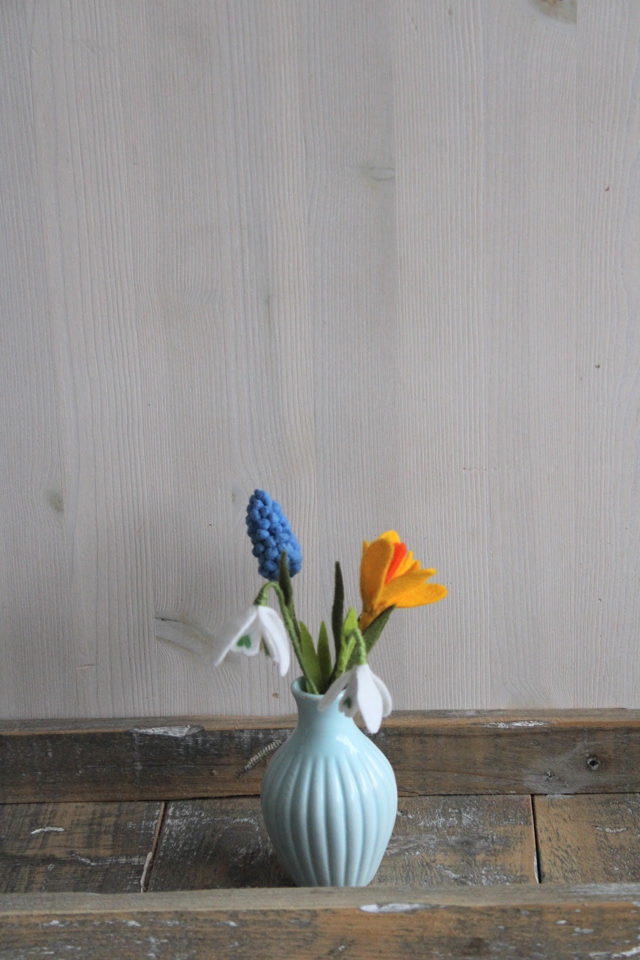 Vilten boeketje met handgemaakte sneeuwklokjes, gele krokus en blauwe druifjes als lente decoratie in een lichtblauwe vaas.