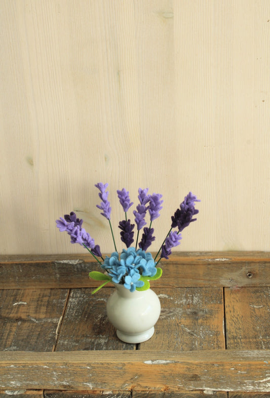 Klein wit vaasje met smalle opening perfect voor handgemaakte viltbloemen zoals deze paarse lavendel en blauwe hortensia op een rustieke houten ondergrond