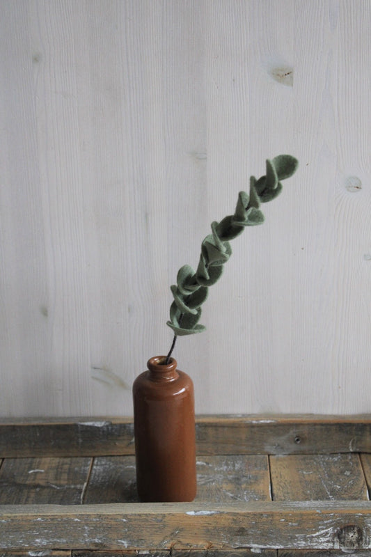 handgemaakte enkele eucalyptus tak van vilt in een bruine keramieken vaas op een houten ondergrond en een lichte achtergrond
