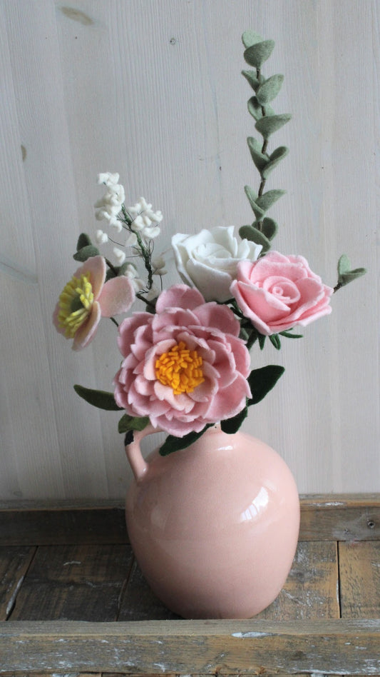 Handgemaakt Boeket van vilten bloemen in roze aardewerk vaas. Het boeket bevat een roze pioenroos, een roze roos, een witte roos, een roze anemoon,  witte gipskruid en eucalyptus takken op houten ondergrond en lichte achtergrond