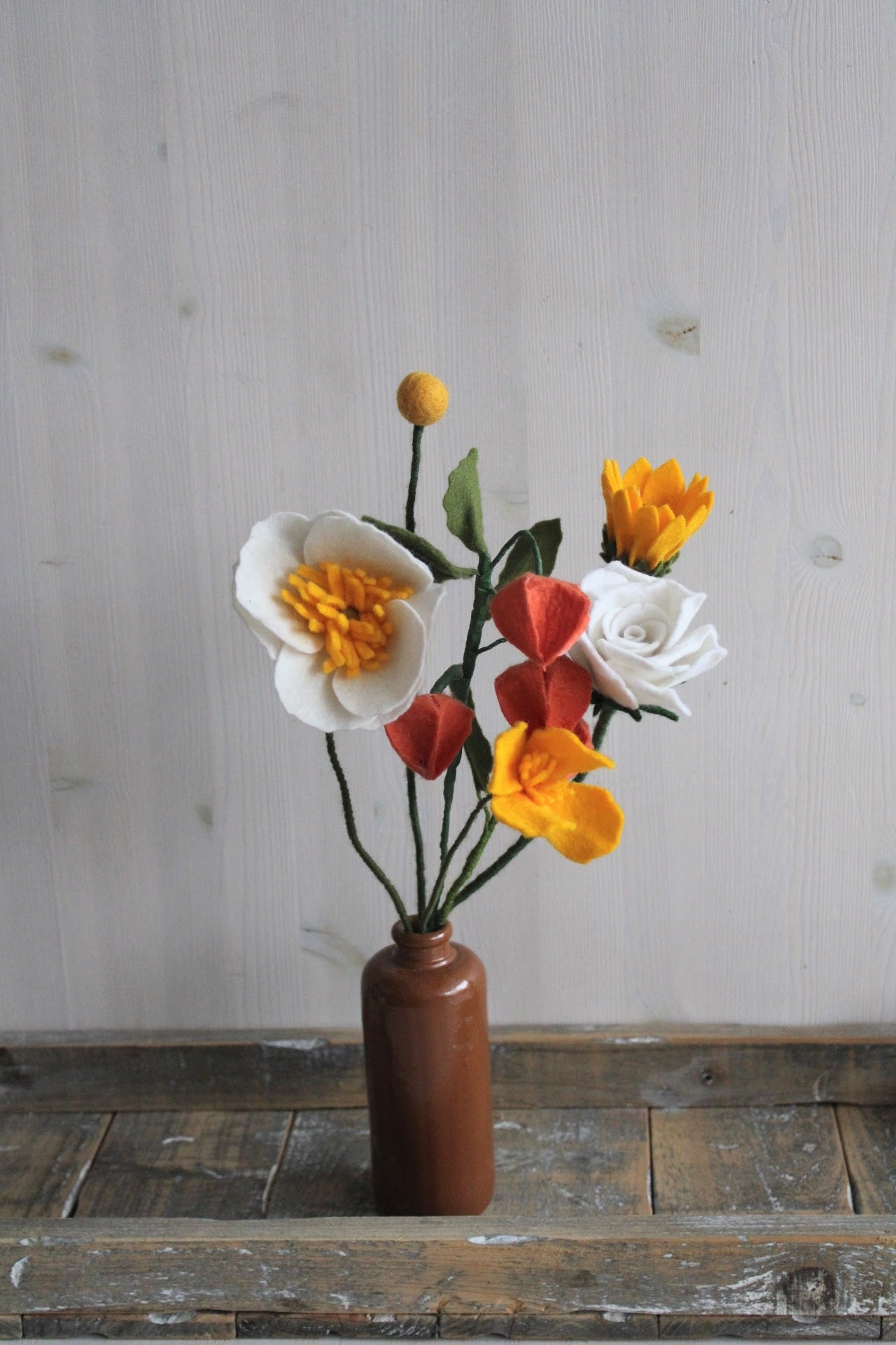 Zomers handgemaakt viltbloemen boeket met oranje lampionnetje, geel slaapmutsje, zonnebloem, witte roos, witte anemoon en gele craspedia in een bruine vaas op een rustikeke houten