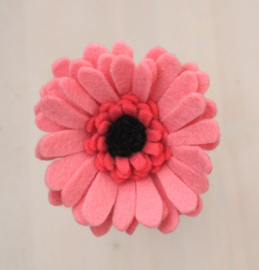 Close-up van een Handgemaakte roze vilten gerbera met een zwart  hartje tegen een lichte achtergrond