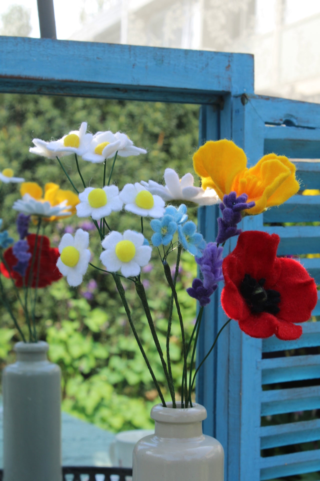 Handgemaakt boeket viltbloemen in een grijze vaas, met madeliefjes, lavendel, vergeet-me-nietjes, gele en rode klaprozen, gepresenteerd voor een blauwe spiegel met tuin op de achtergrond