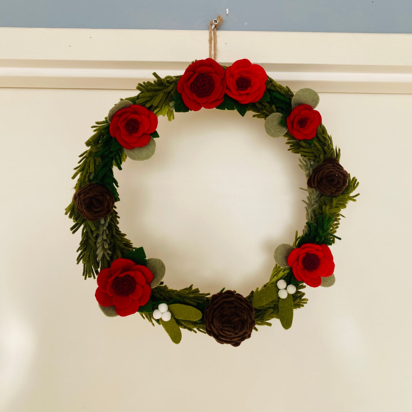 kerstkrans met dennengroen, eucalyptusblad, dennenappels, rode bloemen en misletoe, alles handgemaakt van vilt, hangend op een lichte achtergrond