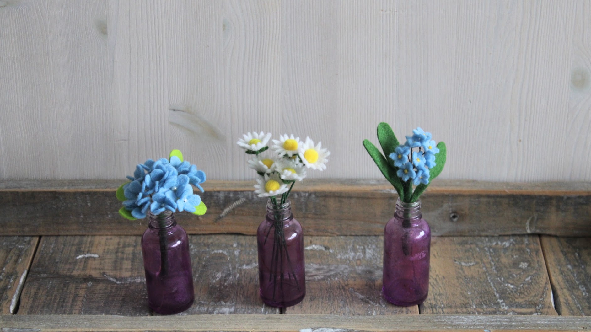Drie mini boeketjes handgemaakt van vilt in paarse glazen flesjes, blauwe hortensia, madeliefjes, blauw vergeet me nietje op een houten rustieke plank