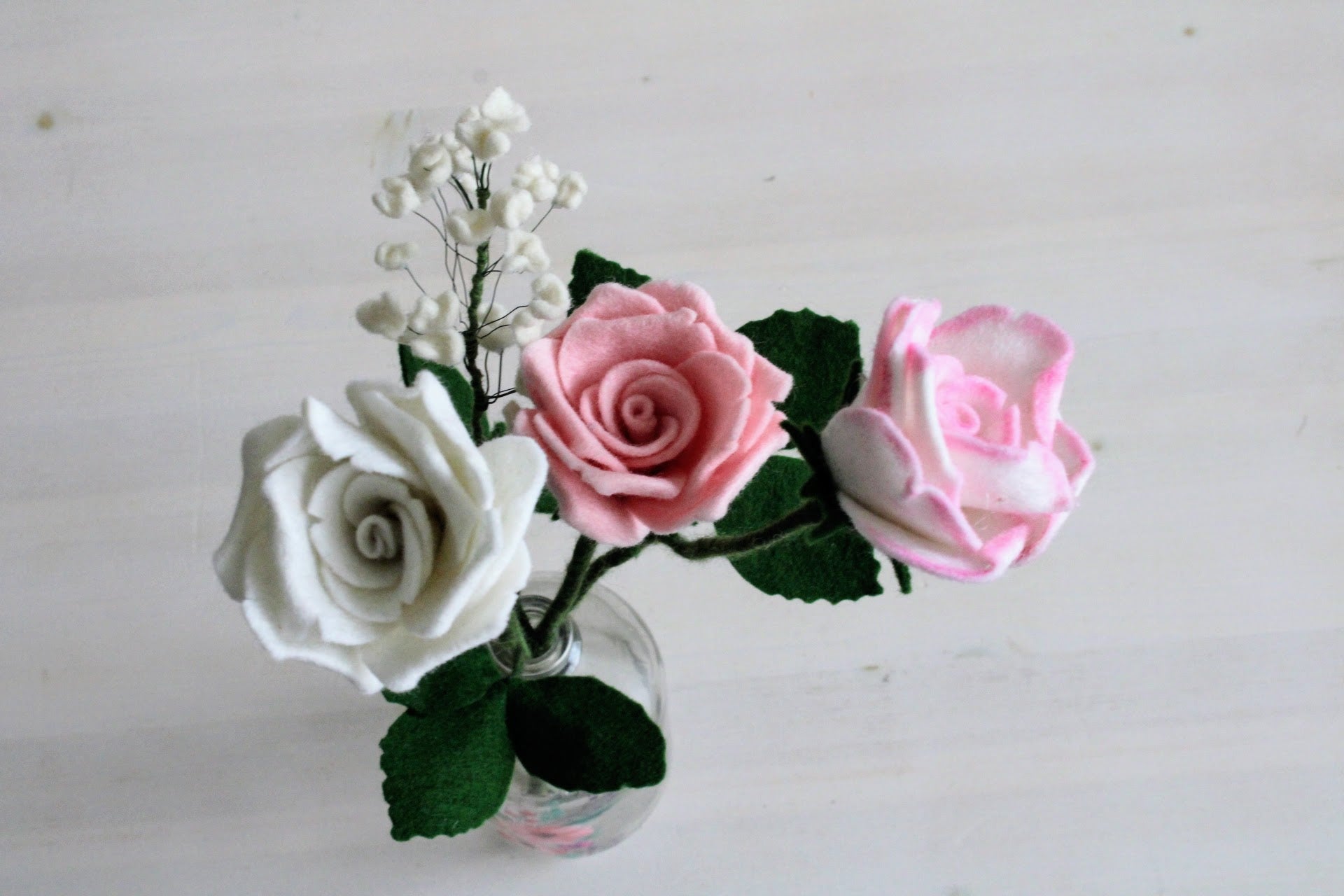 Handgemaakt boeket van viltbloemen in een glazen vaas, met roze en witte rozen en fijn wit gipskruid op een witte ondergrond