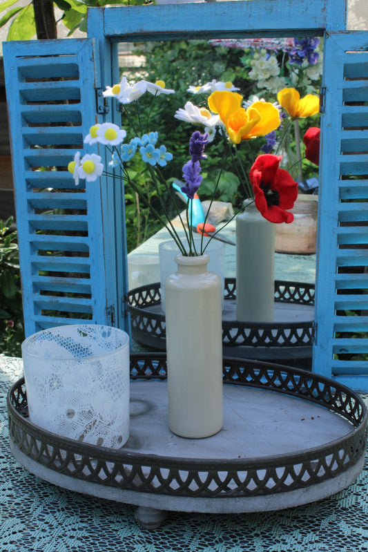 Handgemaakt boeket viltbloemen in een witte vaas, met madeliefjes, lavendel, vergeet-me-nietjes, gele tulpen en rode klaproos, gepresenteerd voor een blauwe spiegel met tuin op de achtergrond